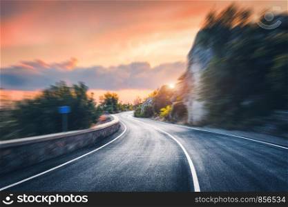 Mountain road at sunset with motion blur effect. Asphalt road and blurred background with rocks, orange sunny sky with clouds in summer. Fast driving. Beautiful highway in motion. Transportation