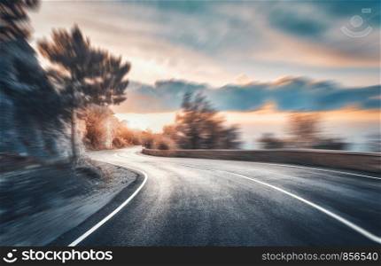 Mountain road at sunset with motion blur effect. Asphalt road and blurred background with rocks, blue sky with sun and clouds in summer. Fast driving. Beautiful highway in motion. Transportation. Mountain road at sunset with motion blur effect. Asphalt road