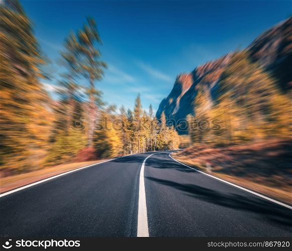 Mountain road in autumn forest at sunset with motion blur effect. Asphalt road and blurred background with orange trees, blue sky with sun in fall. Fast driving. Beautiful highway. Transportation. Mountain road in autumn forest at sunset with motion blur effect