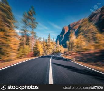 Mountain road in autumn forest at sunset with motion blur effect. Asphalt road and blurred background with orange trees, blue sky with sun in fall. Fast driving. Beautiful highway. Transportation. Mountain road in autumn forest at sunset with motion blur effect