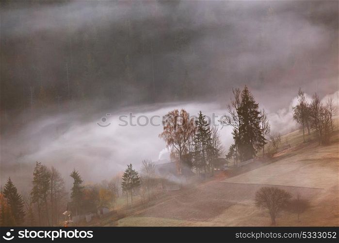 Mountain village in clouds of fog and smoke in the autumn morning. Farm on hills in the mountains. Waves of mist in a forest.