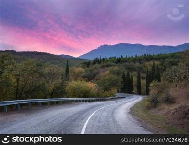 Mountain winding road passing through the forest with colorful sky and pink clouds at sunset in summer 