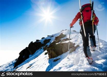 Mountaineer walking up along a steep snowy ridge with the skis in the backpack. In background a dramatic sky with a shiny bright sun. Concepts: adventure, achievement, courage, determination, self-realization, dangerous activity, extreme sport, winter leisure.