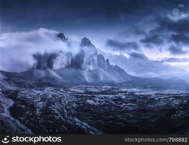 Mountains in fog at beautiful night. Dreamy landscape with mountain peaks, stones, grass, purple sky with blurred low clouds, stars and moon. Rocks at dusk. Tre Cime in Dolomites, Italy. Italian alps. Mountains in fog at beautiful night. Dreamy landscape