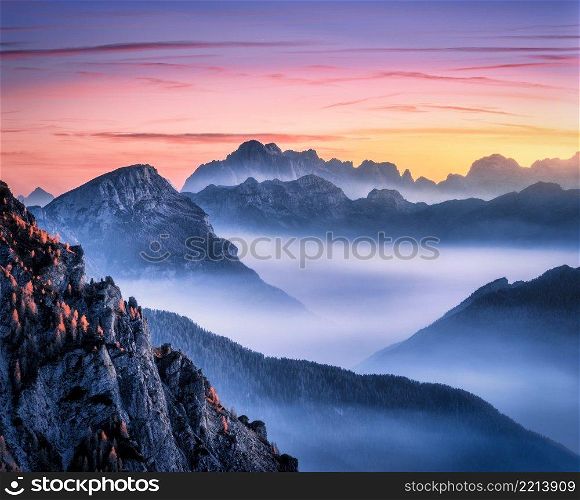 Mountains in fog at beautiful sunset in autumn in Dolomites, Italy. Landscape with alpine mountain valley, low clouds, trees on hills, colorful sky at dusk. Aerial view. Passo Giau at night. Nature