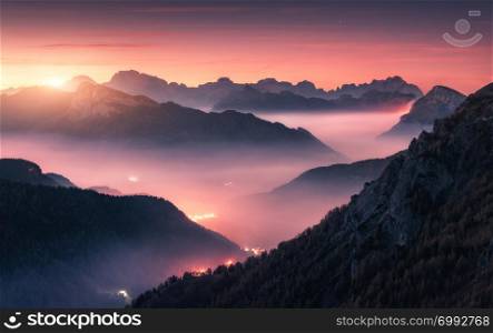 Mountains in fog at beautiful sunset in autumn in Dolomites, Italy. Landscape with alpine mountain valley, low clouds, forest, purple sky city illumination at twilight. Aerial view. Passo Giau