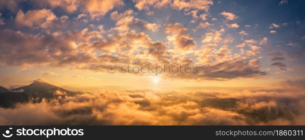 Mountains in low clouds at sunrise in summer. Aerial view of mountain peak in fog. Beautiful landscape with high rocks, forest, colorful sky. View from above of mountain valley in clouds. Foggy hills
