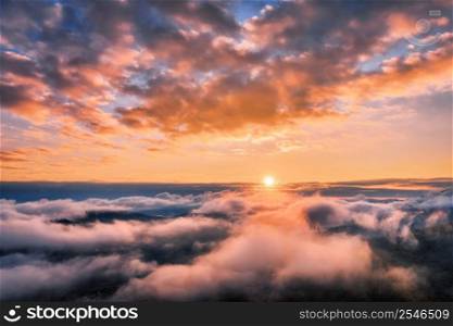 Mountains in low clouds at sunset in summer. Aerial view of mountain hills in fog. Beautiful landscape with high rocks, forest, colorful sky. View from above of mountain valley in clouds. Foggy hills