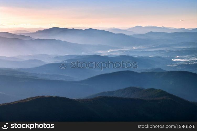 Mountains lighted by bright sunbeams at sunset in autumn. Landscape with mountain ridges in fog, rocks, forest, colorful sky with orange sunlight in fall. Aerial view of mountain valley. Top view