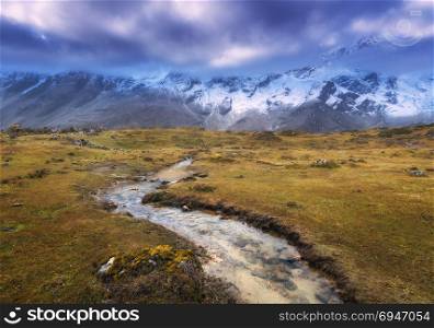 Mountains with snow covered peaks, small river, yellow grass and cloudy sky at sunset. Mountain valley. Adventure in Nepal. Amazing scene with Himalayan mountains. Beautiful landscape. Nature. Travel. Mountains with snow covered peaks, small river, yellow grass