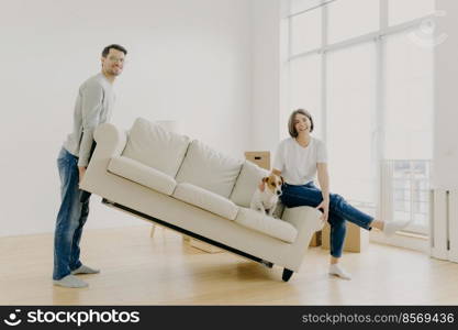Move into new home. Husband and wife carry sofa, furnish living room after renovation, happy to buy apartment, lovely pet poses on couch, work together as team, place furniture in empty room