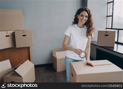 Mover is packing cargo with duct tape. Young lady is wrapping cardboard boxes with packing tape. Happy hispanic girl relocates. Moving service worker preparing boxes for shipment.. Mover is packing cargo with duct tape. Young lady is wrapping cardboard boxes with packing tape.