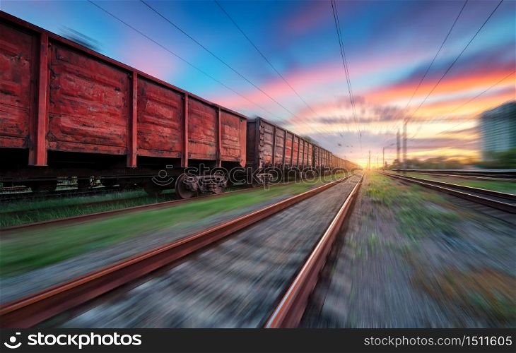 Moving freight train at sunset. Railroad and beautiful sky with clouds with motion blur effect in summer. Industrial landscape with train, railway station and blurred background. Railway platform
