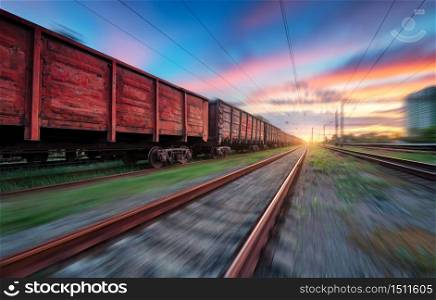Moving freight train at sunset. Railroad and beautiful sky with clouds with motion blur effect in summer. Industrial landscape with train, railway station and blurred background. Railway platform