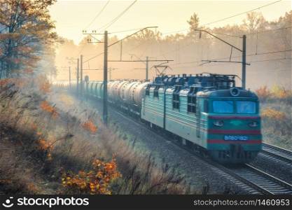 Moving freight train in beautiful forest in fog at sunrise in autumn. Colorful landscape with train in motion, railroad, gold sunbeams, foggy trees, orange sky in fall. Railway station. Transportation