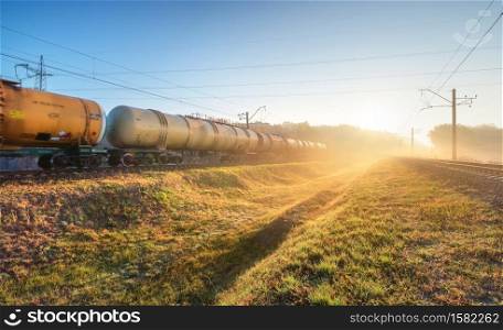 Moving freight train in fog at sunrise in autumn. Industrial landscape with train in motion, railroad, green grass, trees, gold sunbeams, foggy trees, blue sky in fall. Railway station. Transportation. Moving freight train in fog at sunrise in autumn. Industrial