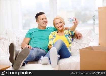moving, home and couple concept - smiling couple relaxing on sofa taking picture with digital camera in new home