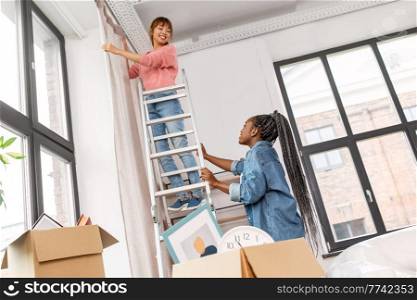 moving, home improvement and real estate concept - happy smiling women with ladder hanging curtain. woman on ladder hanging curtains at home