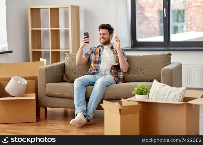 moving, people and real estate concept - happy man with smartphone and boxes sitting on sofa and having video call at new home. man with smartphone having video call at new home