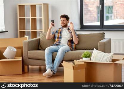 moving, people and real estate concept - happy man with smartphone and boxes sitting on sofa and having video call at new home. man with smartphone having video call at new home