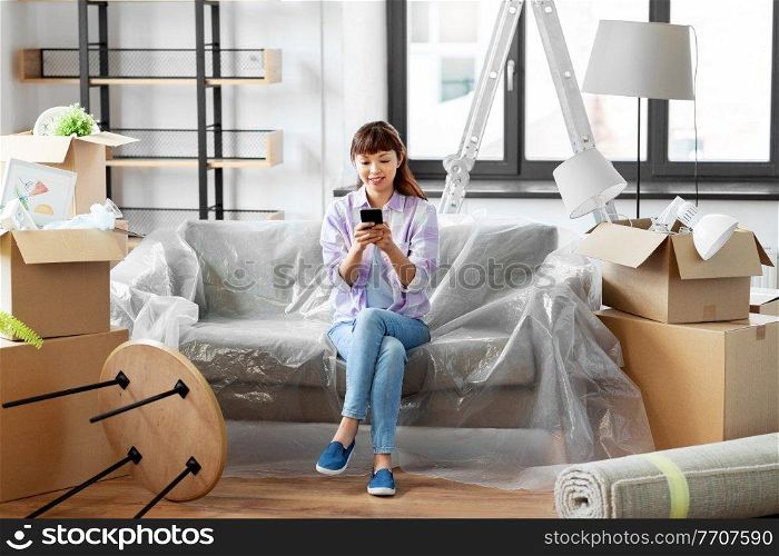 moving, people and real estate concept - happy smiling asian woman with smartphone and boxes at new home. asian woman with smartphone moving into new home