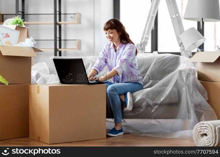 moving, people and real estate concept - happy smiling asian woman with laptop computer and boxes at new home. woman with laptop moving into new home