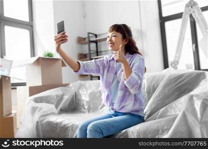 moving, people and real estate concept - happy smiling asian woman with smartphone and boxes having video call at new home and showing thumbs up. woman with phone having video call at new home