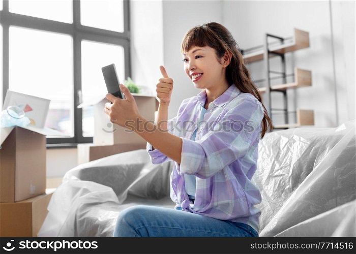 moving, people and real estate concept - happy smiling asian woman with smartphone and boxes having video call at new home and showing thumbs up. woman with phone having video call at new home