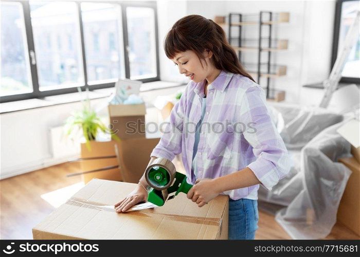 moving, people and real estate concept - happy smiling asian woman with adhesive tape packing box at new home. woman with adhesive tape packing box at new home