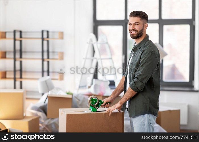 moving, people and real estate concept - happy smiling man with adhesive tape packing box at new home. man with adhesive tape packing box at new home