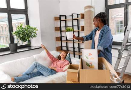 moving, people and real estate concept - happy smiling woman unpacking box and her friend resting on sofa at new home. woman unpacking boxes and moving to new home