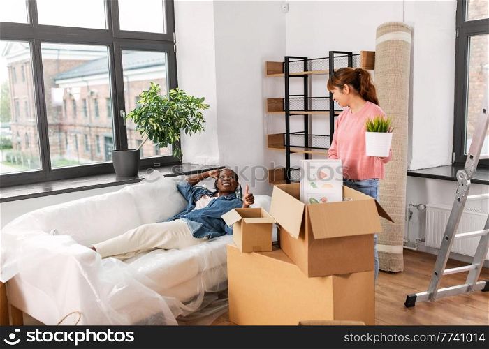 moving, people and real estate concept - happy smiling woman unpacking box and her friend resting on sofa at new home. woman unpacking boxes and moving to new home