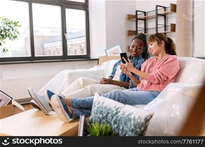 moving, people and real estate concept - happy smiling women with smartphones and boxes at new home. women with smartphones moving into new home