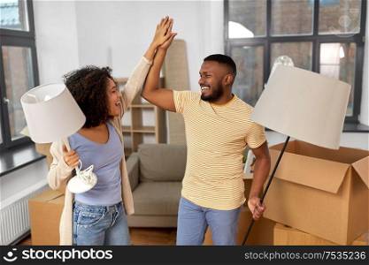 moving, people, repair and real estate concept - happy african american couple with lamps packing stuff into cardboard boxes at home and making high five. happy couple packing boxes and moving to new home