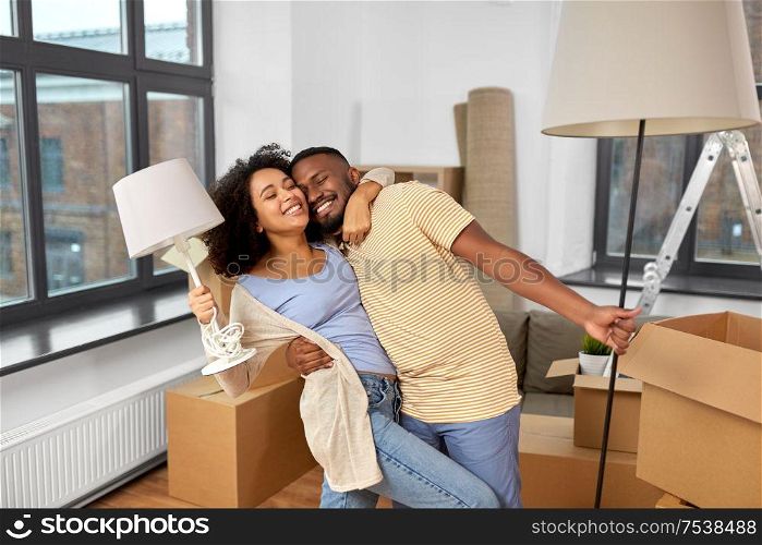 moving, people, repair and real estate concept - happy african american couple with lamps packing stuff into cardboard boxes at home and hugging. happy couple packing boxes and moving to new home