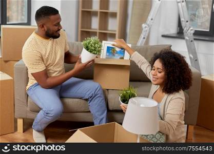 moving, people, repair and real estate concept - happy african american couple with photo frame and cardboard boxes sitting on sofa at new home. happy couple packing boxes and moving to new home