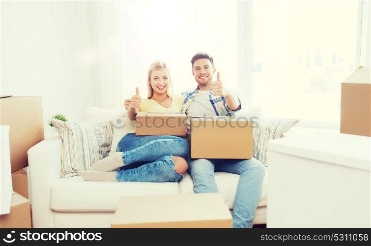 moving, people, repair and real estate concept - happy couple with big cardboard boxes showing thumbs up on sofa at new home. happy couple with boxes showing thumbs up at home