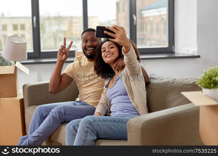 moving, repair and real estate concept - happy african american couple with cardboard boxes sitting on sofa and taking selfie by smartphone at new home. happy couple moving to new home and taking selfie