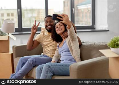 moving, repair and real estate concept - happy african american couple with cardboard boxes sitting on sofa and taking selfie by smartphone at new home. happy couple moving to new home and taking selfie