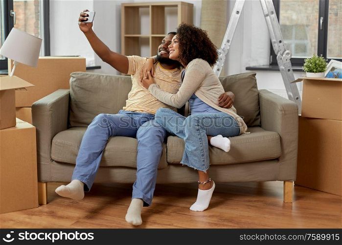 moving, repair and real estate concept - happy african american couple with cardboard boxes sitting on sofa and taking selfie by smartphone at new home. happy couple moving to new home and taking selfie