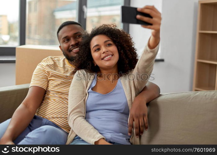 moving, repair and real estate concept - happy african american couple with cardboard boxes sitting on sofa and taking selfie by smartphone at new home. happy couple moving to new home and taking selfie