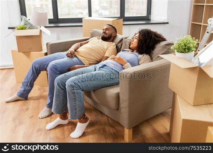 moving, repair and real estate concept - tired african american couple with cardboard boxes relaxing on sofa at new home. couple with boxes tired after moving to new home