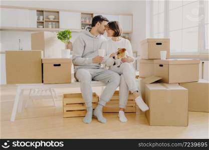 Moving to new home, family time concept. Couple in love going to kiss each other, sit at white bench against kitchen interior, play with dog, surrounded with many packages, enjoy first day in new home