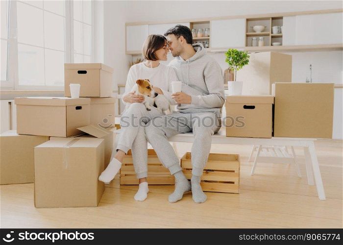 Moving to new home, family time concept. Couple in love going to kiss each other, sit at white bench against kitchen interior, play with dog, surrounded with many packages, enjoy first day in new home