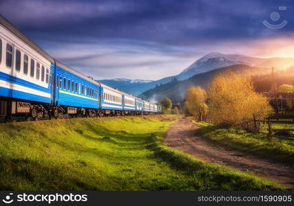 Moving train in mountains at sunset in autumn. Industrial landscape with passenger speed train on railroad, dirt road, snowy rocks, orange trees, green grass, purple sky in fall. Rural railway station