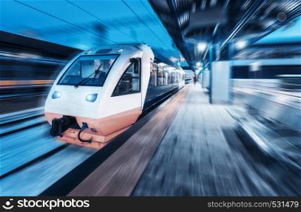 Moving train on the railway station at night with motion blur effect. Urban landscape with modern high speed train on the railway platform with illumination. Intercity vehicle. Passenger railroad