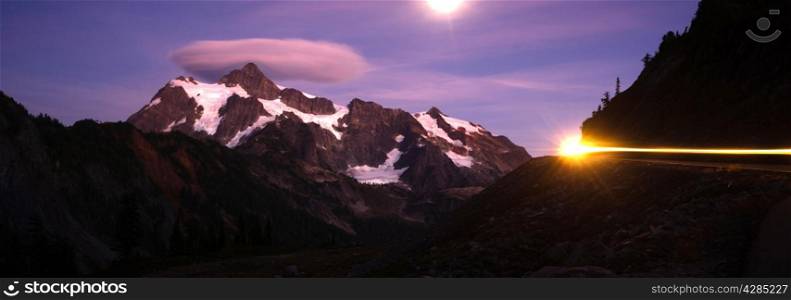 Mt Shuksan is lighted by a full moon as a lone car passes on the roadway in a long exposure