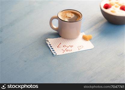 Mug of Arabic coffee and a heart shaped sugar on a math paper with the love you message and a bowl of yogurt on a blue wooden table, in the morning light.