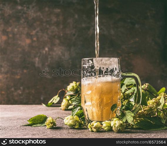 Mug of beer on a rustic table with a vine and cones of hops opposite a dark wall, front view. Beer is poured into a mug