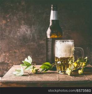 Mug of beer with foam and cold bottle of beer with dew drops and hops vines on a rustic table opposite a dark wall background, front view